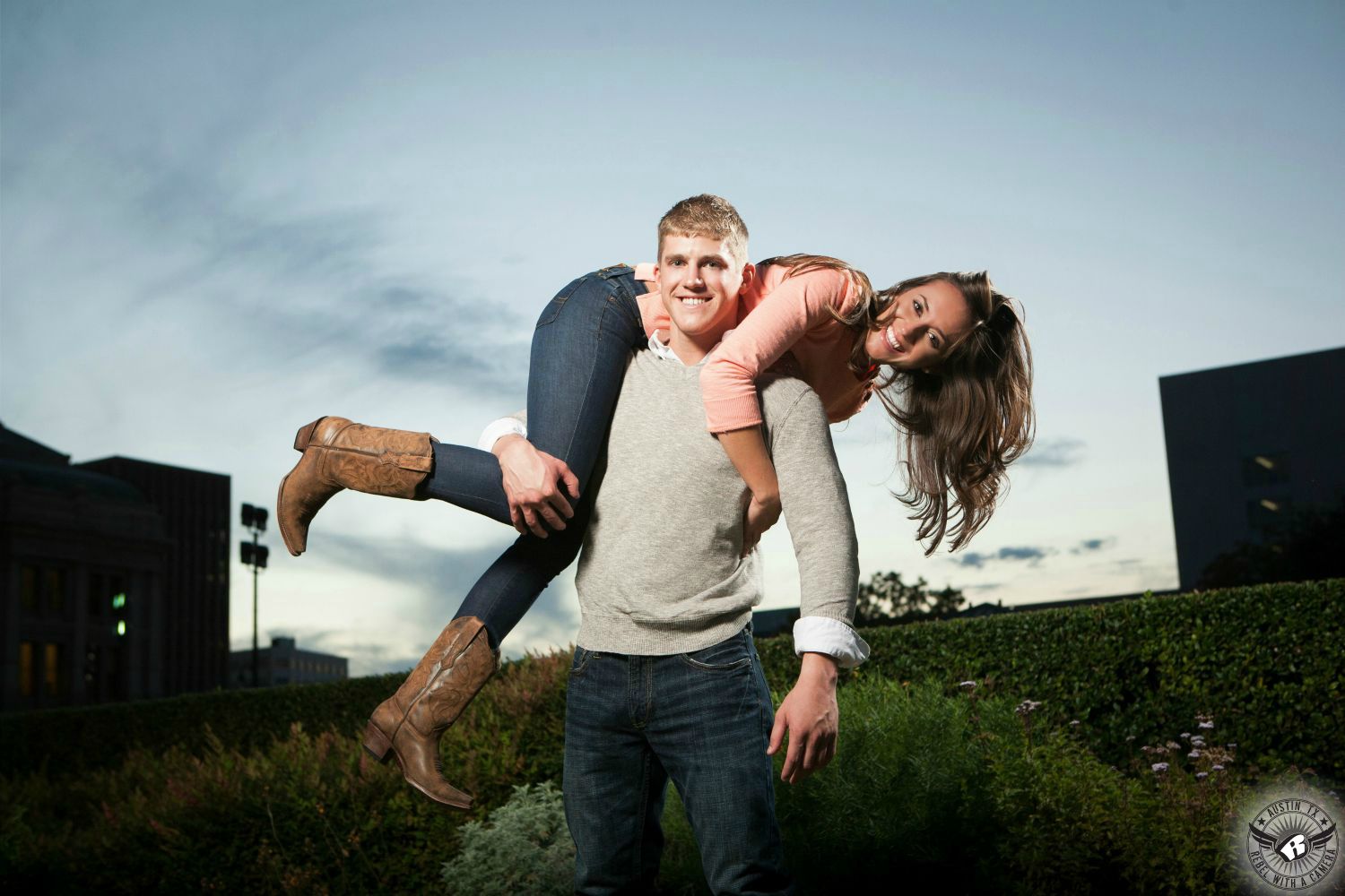 Smiling, sandy short crew cut haired guy with a grey sweater with rolled up sleeves and blue jeans throws brown haired girl wearing a pink long sleeve shirt, blue jeans and brown cowboy boots over shoulder behind the Texas Capital with bushes and hedges visible in this happy engagement photograph in Austin.